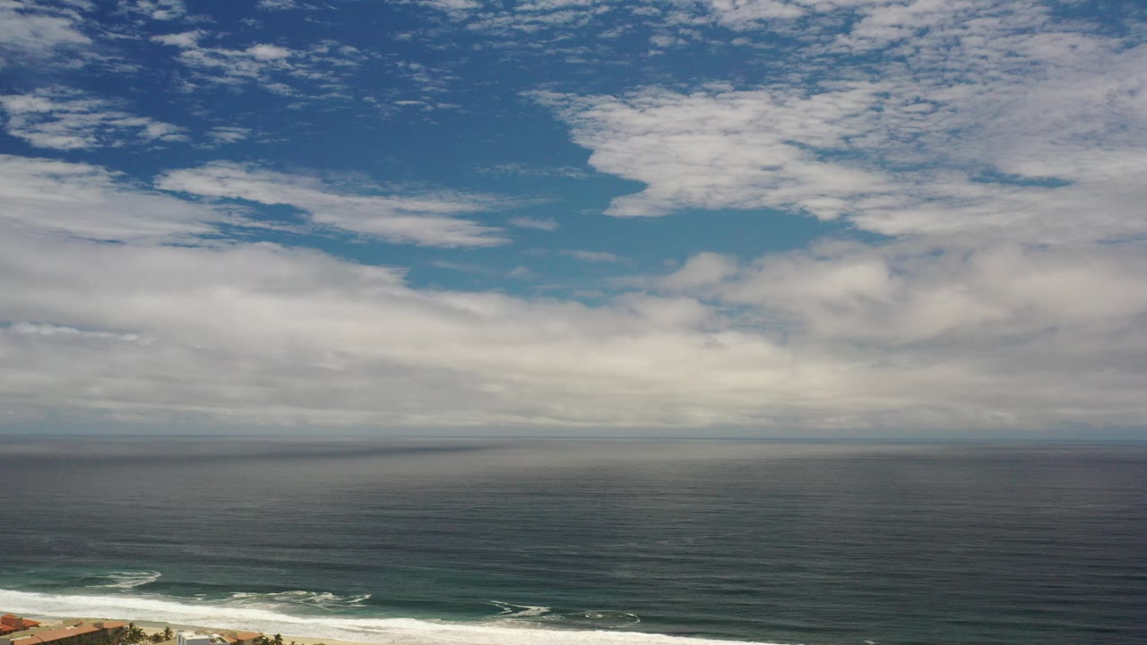 Opening frame of a Los Cabos, Mexico ocean aerial with blue-green water, a level horizon, broken white clouds, surf at the lower edge, and no visible people.