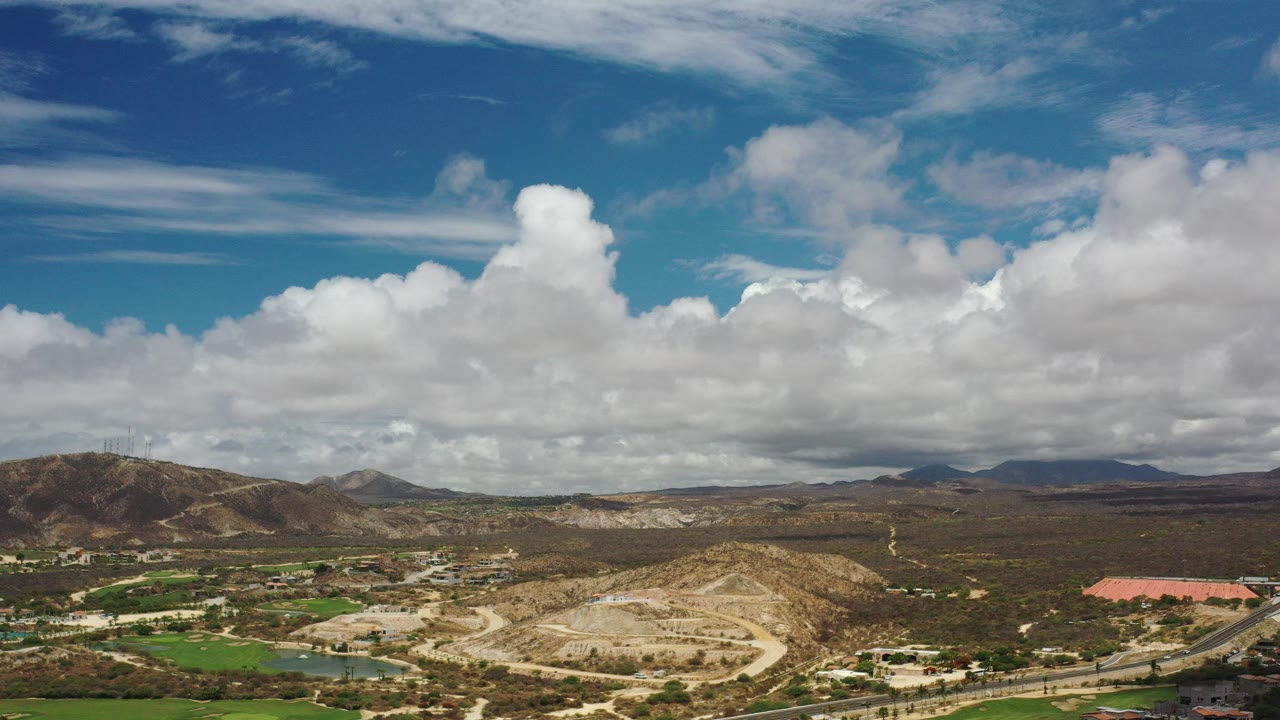 Wide aerial view of clouds over dry hills, roads, green grounds, and distant mountains.