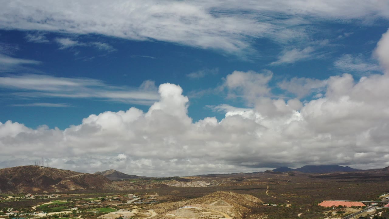 Opening wide view in Los Cabos, Mexico, with dry hills, distant mountains, scattered greenery, blue sky, layered white clouds, and no visible people.