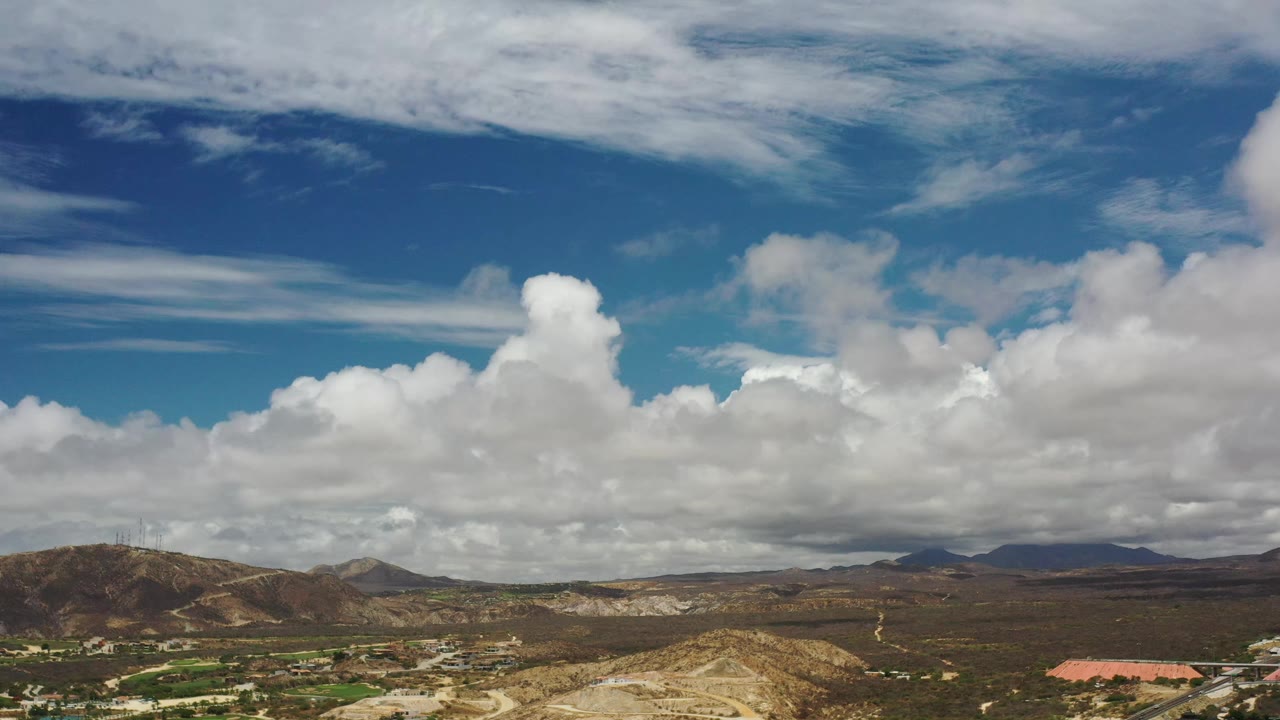 Wide Los Cabos landscape with brown rolling hills, a darker mountain line on the horizon, bright cloud masses, and no visible people.