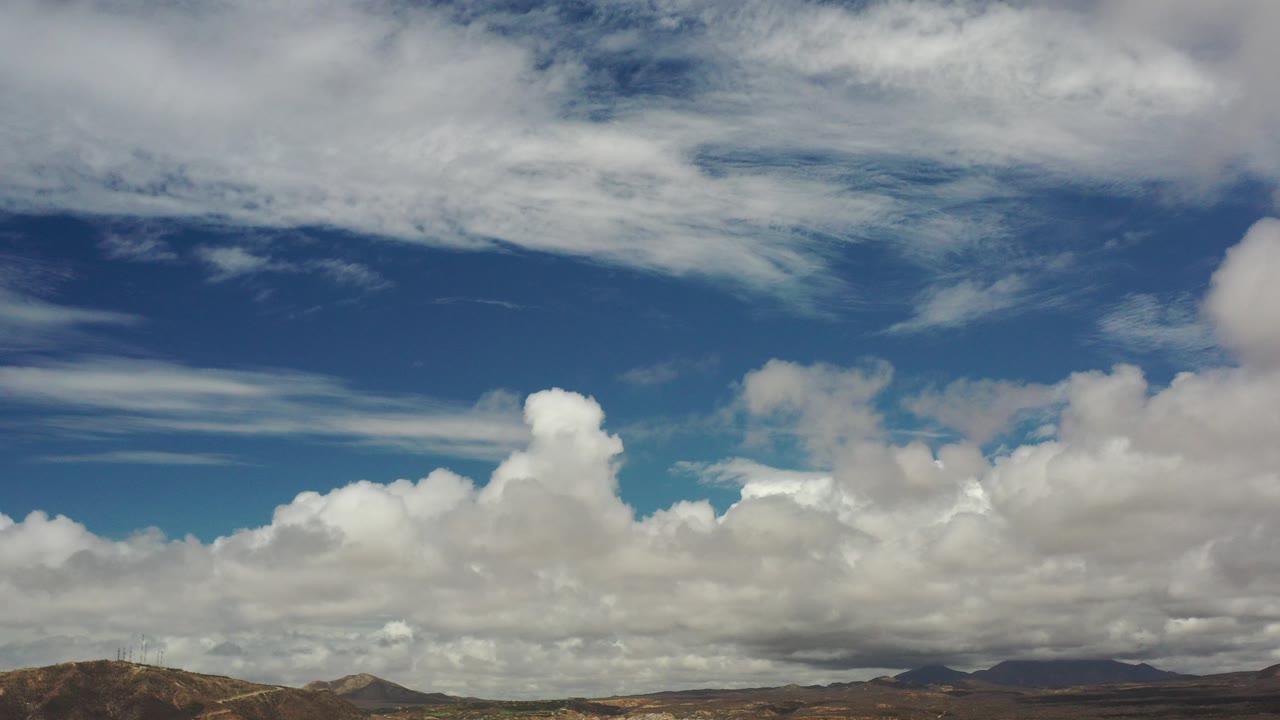 Opening frame of a Los Cabos, Mexico sky view with broad white clouds, blue patches, a narrow line of brown hills, and no visible people.
