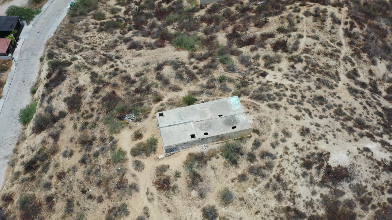 Aerial frame in Los Cabos, Mexico with the rectangular concrete building centered lower in sandy terrain, sparse shrubs around it, a road at the left side, and no visible people.