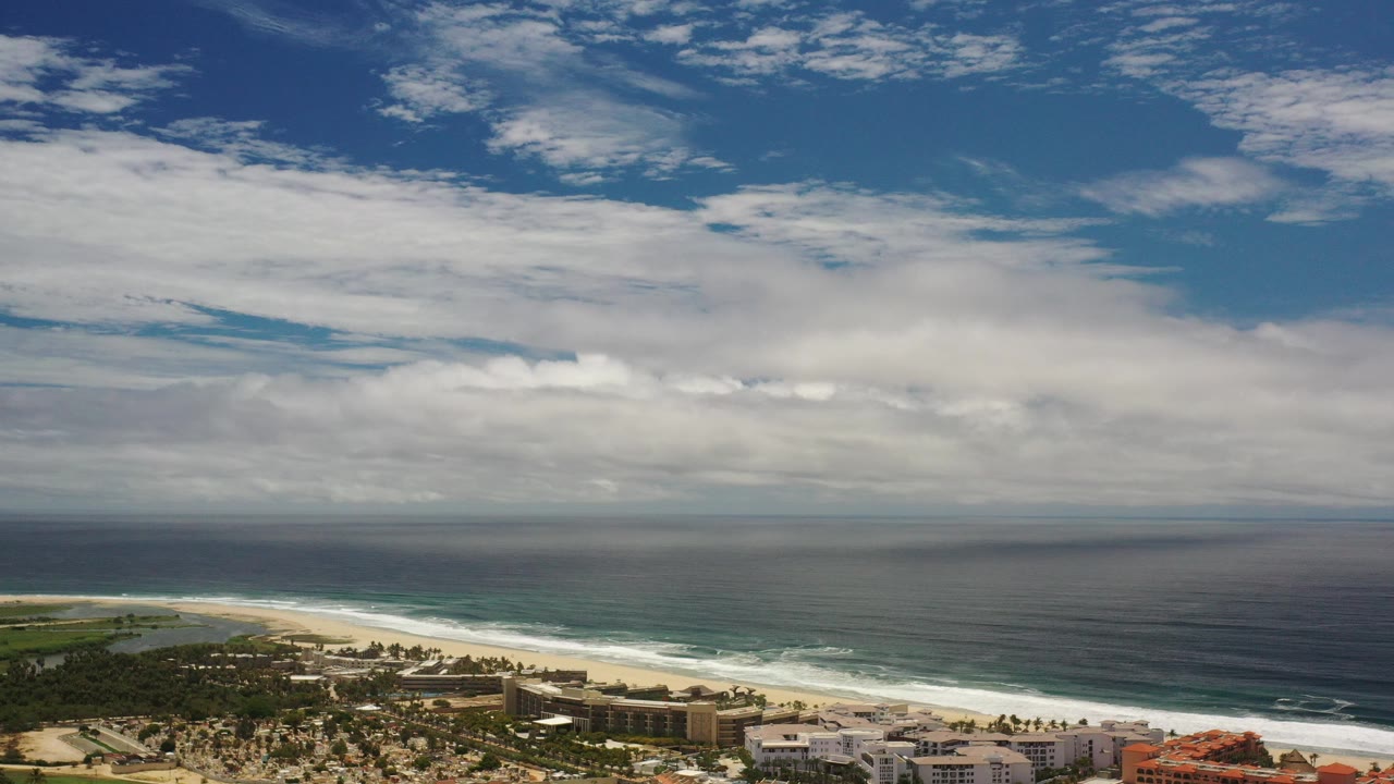 Aerial coastline with ocean, beach, buildings, roads, and clouded sky.
