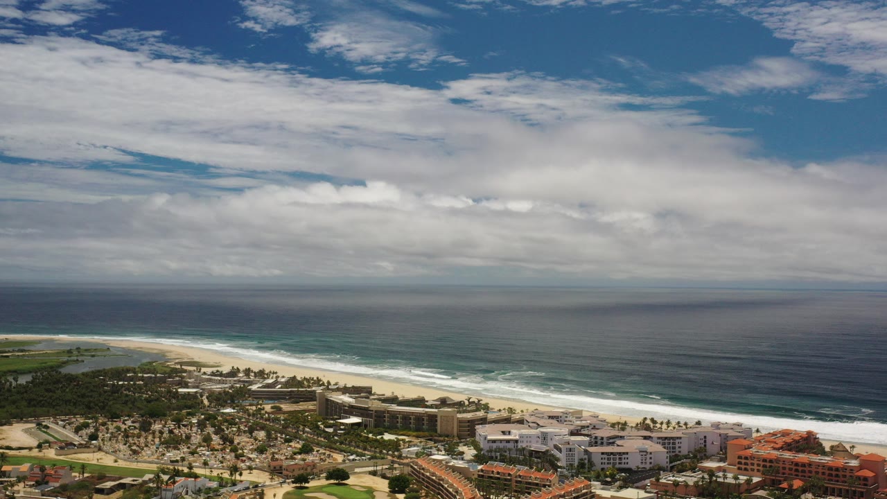 Opening frame of Los Cabos coastline with deep blue ocean, white surf, sandy beach, shoreline buildings, green land, broad cloud cover, and no visible people.