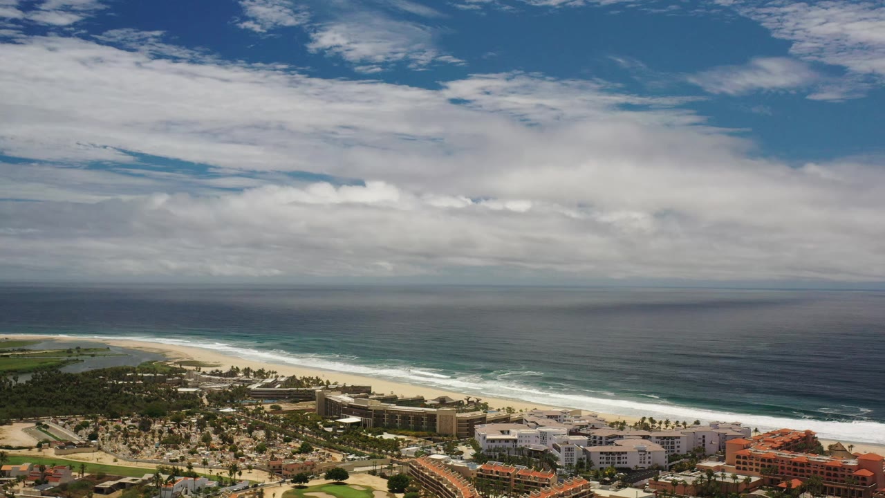 Los Cabos aerial frame with the ocean horizon across the center, pale beach and low shoreline buildings below, white cloud bands above, and no visible people.