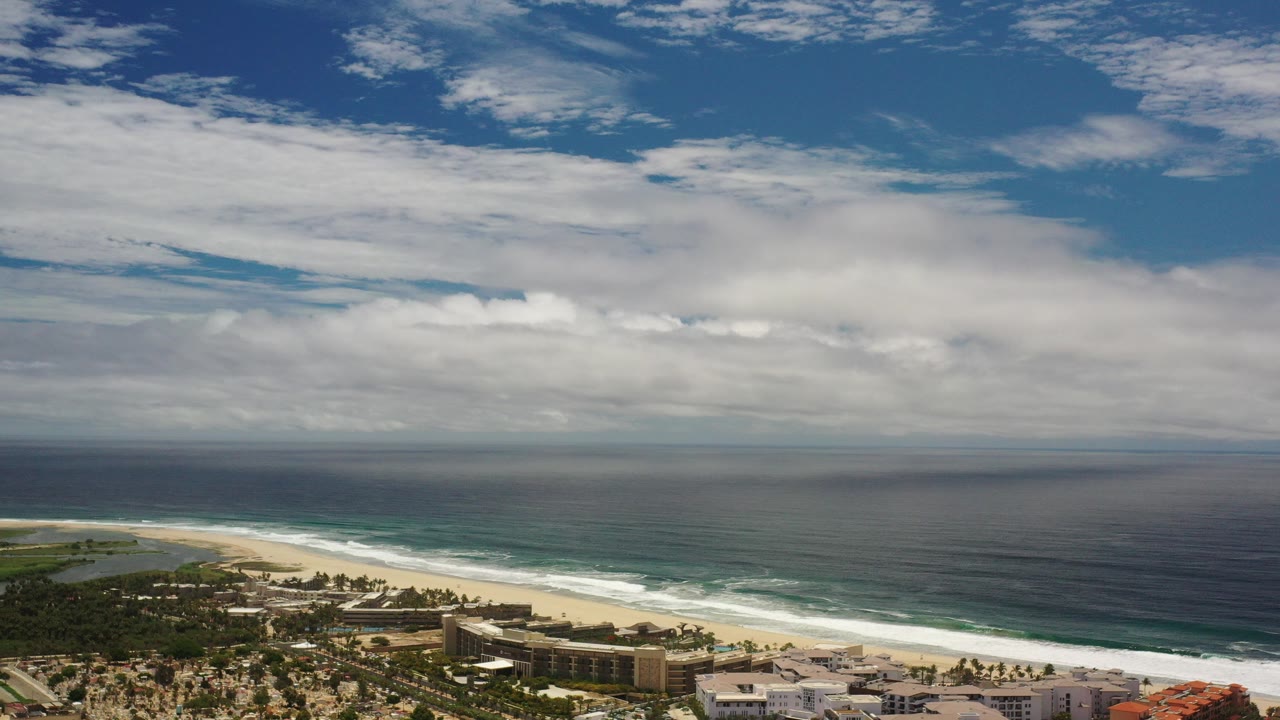 Opening aerial view over the Los Cabos coastline, with dark blue ocean patches, white surf, sandy beach, low buildings, and no visible people.