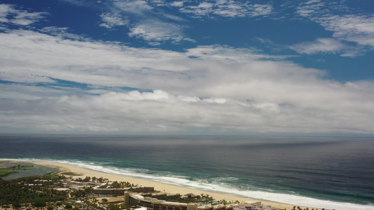 Opening frame of a Los Cabos coastal view with sandy beach, white surf, dark ocean water, low buildings at the bottom edge, layered clouds, and no visible people.