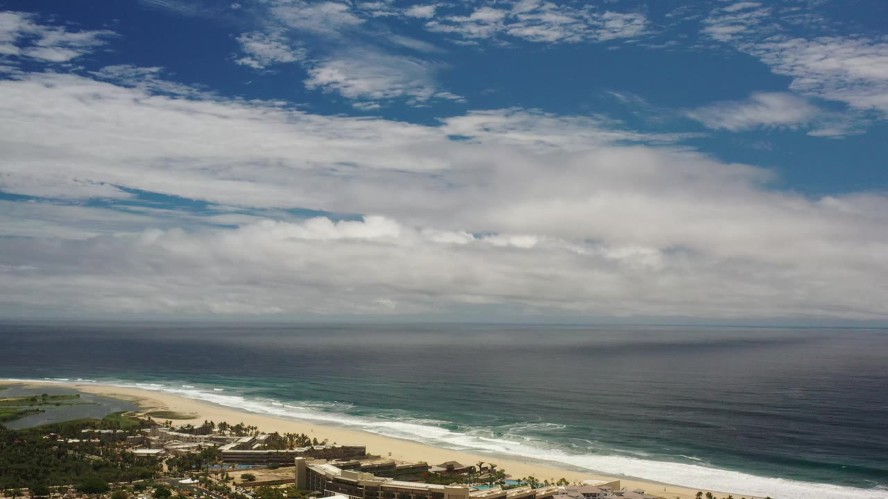 Early frame from the Los Cabos view showing the shoreline angled across the lower frame, ocean filling the middle, a cloudy horizon, and no visible people.