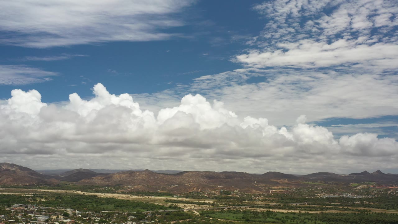 Wide aerial view of clouds, mountains, greenery, and scattered buildings.