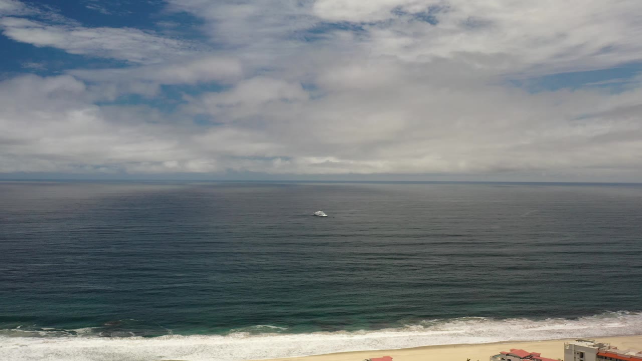 Opening view of a small gray ship offshore in Los Cabos, Mexico, with green-blue water, a narrow band of white surf, cloudy sky, and no visible people.