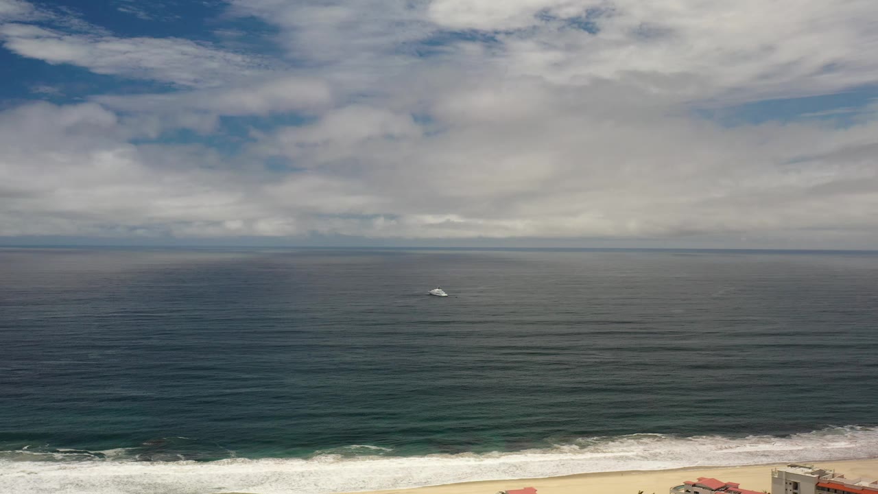 A small gray ship remains near the center of the Los Cabos ocean view, with the white shoreline break lower in frame and no visible people.