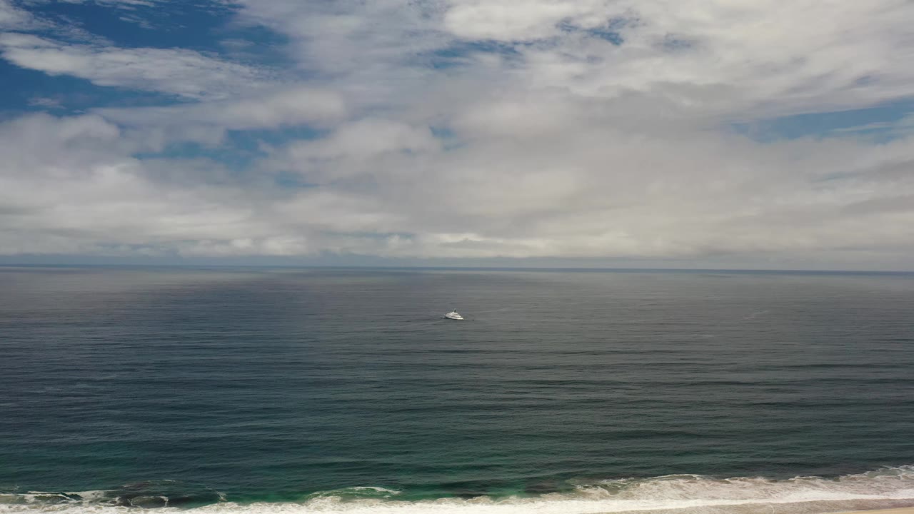 Wide Los Cabos ocean frame with a small white boat left of center, layered cloud cover, muted blue-green water, and no visible people.