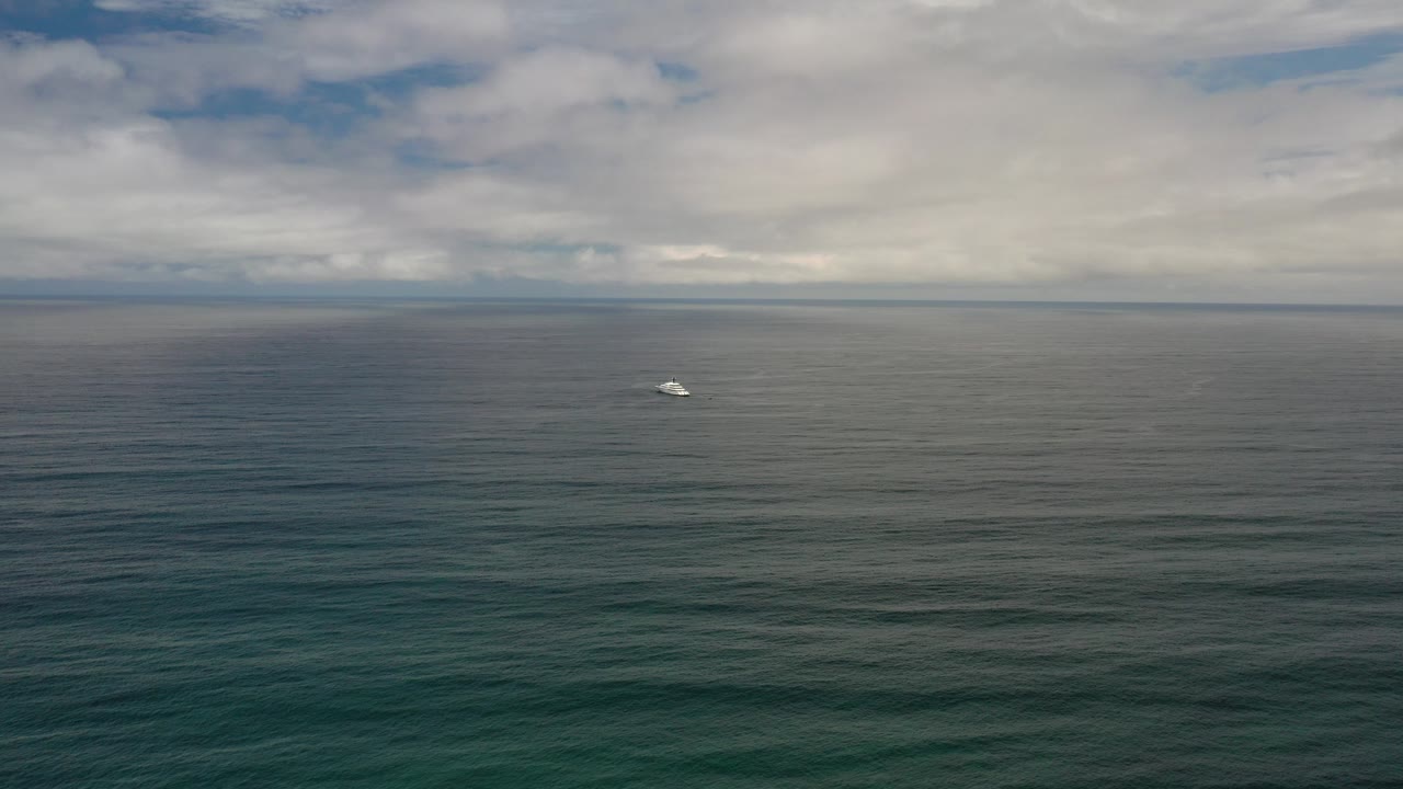 Opening view of the Los Cabos ocean with teal water in the foreground, a small white boat near the left side of the distant horizon, gray clouds overhead, and no visible people.