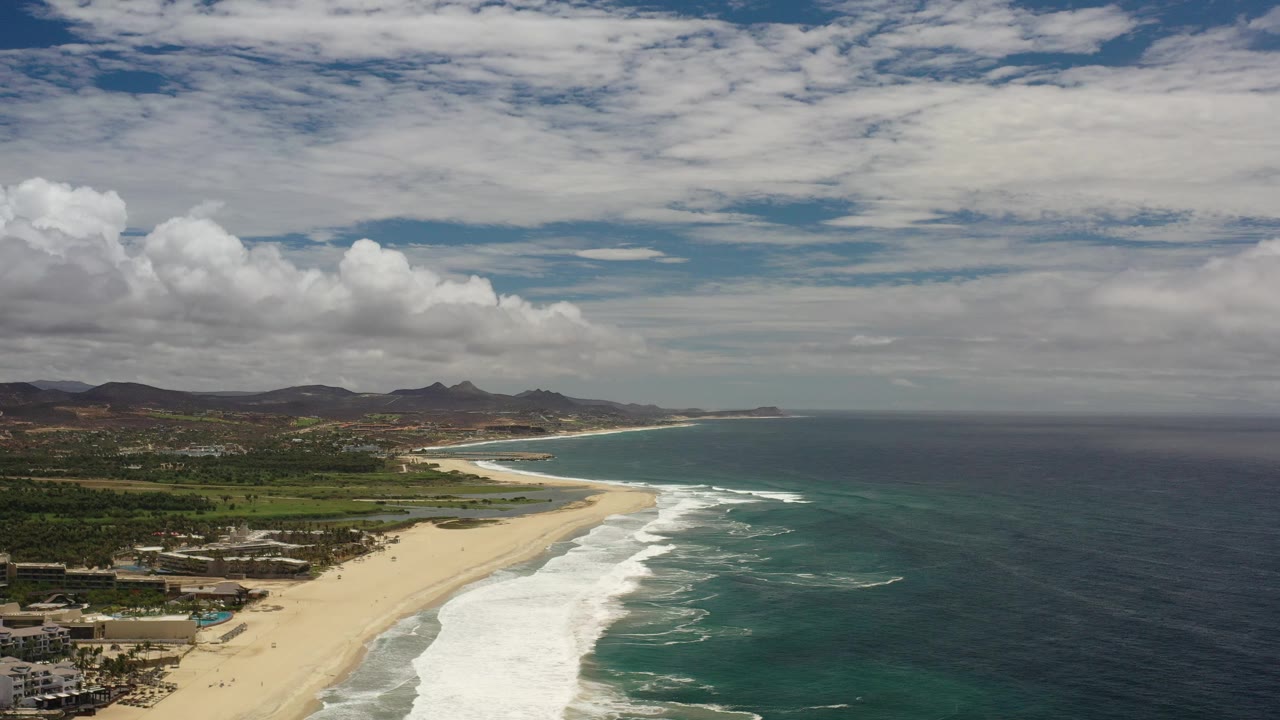 Wide opening frame of the Los Cabos, Mexico coastline with a sandy beach at left, white surf lines, blue-green ocean at right, distant hills, layered clouds, and no visible people.