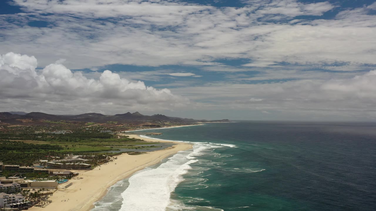 Los Cabos, Mexico aerial view one second in, showing white surf spreading along the sand, darker blue water offshore, coastal buildings near the lower left, hills on the horizon, and no visible people.