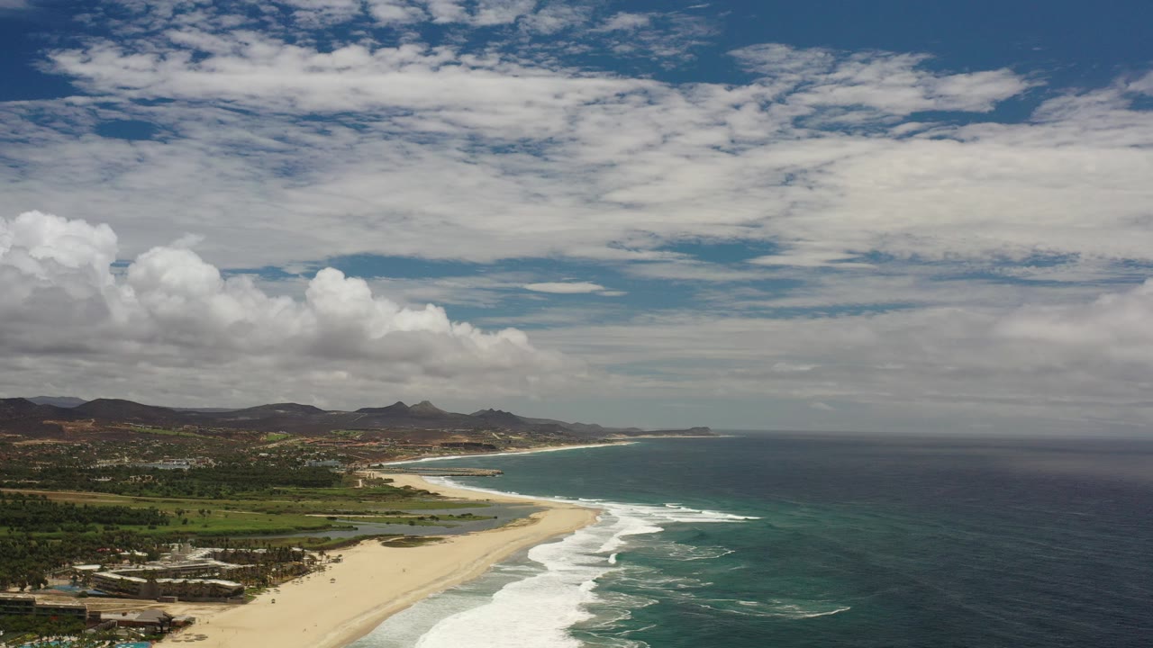 Aerial coastline with sandy beach, surf, ocean, hills, and clouds.