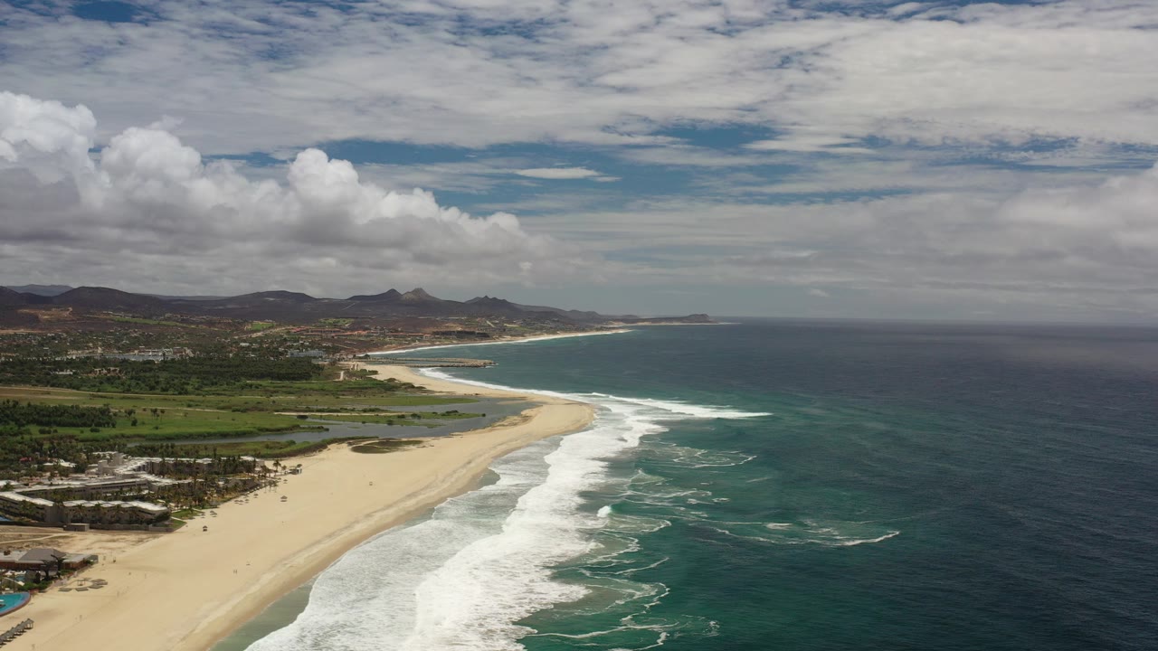 Aerial view over a Los Cabos, Mexico beach with white surf rolling toward sand, green dune vegetation inland, distant hills, and no visible people.