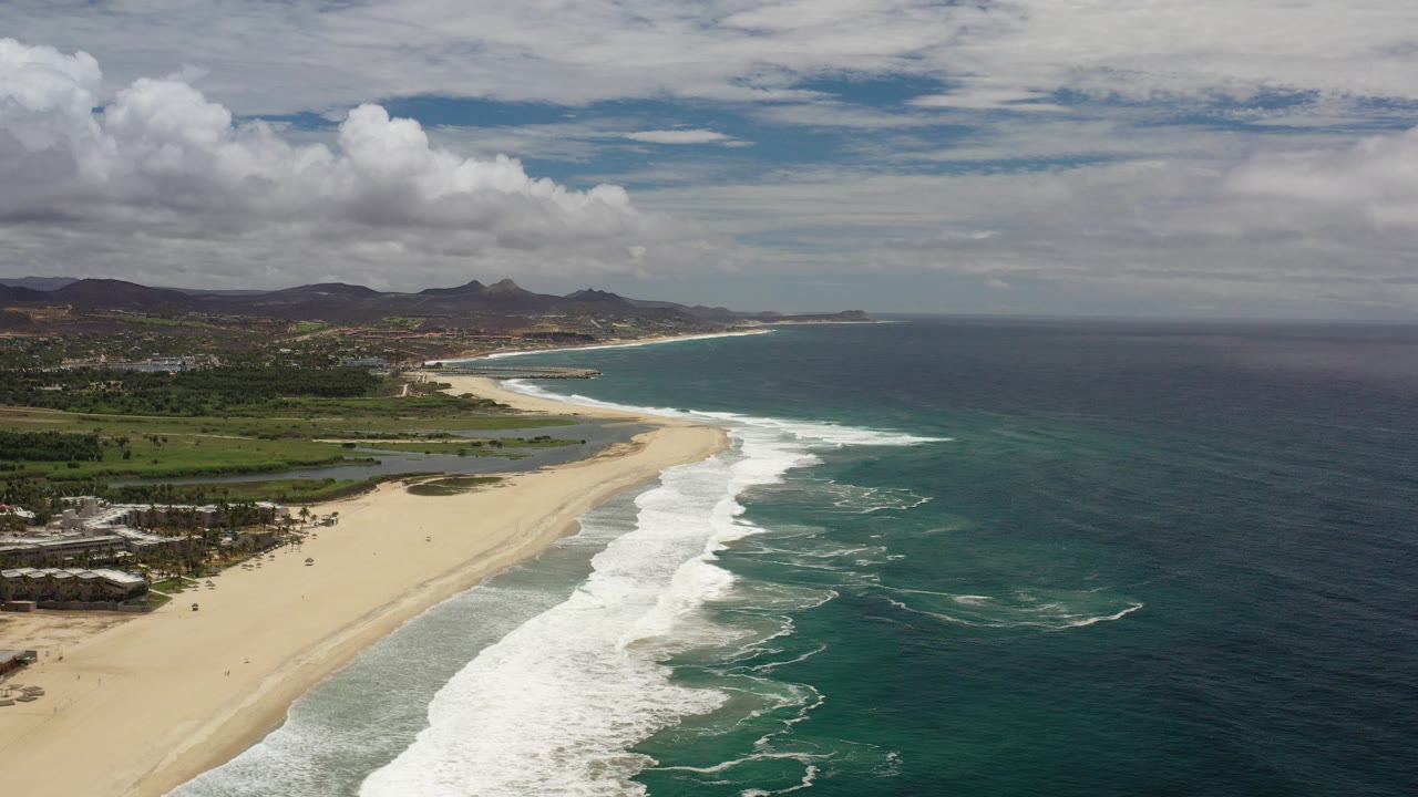 Aerial coastline with sandy beach, surf, ocean, inland greenery, and hills.