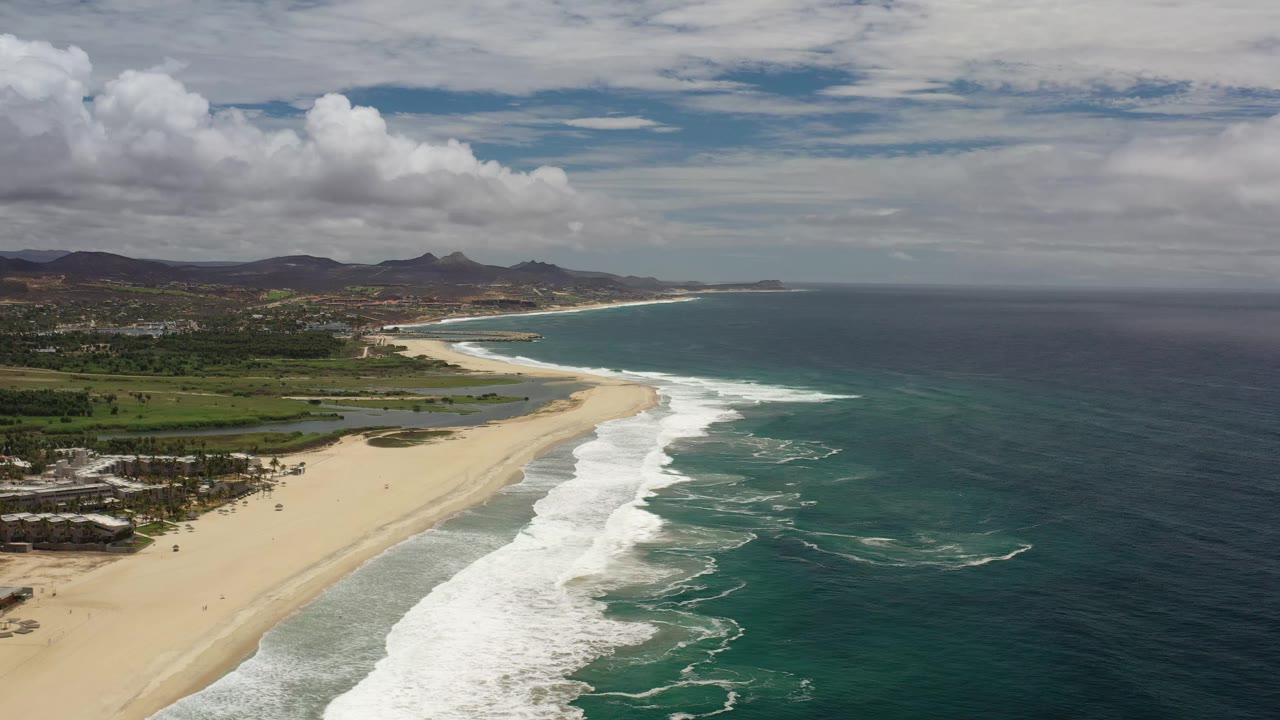 The Los Cabos aerial frame holds a wide diagonal shoreline, with foamy waves crossing the sand, blue-green water to the right, low hills beyond, and no visible people.