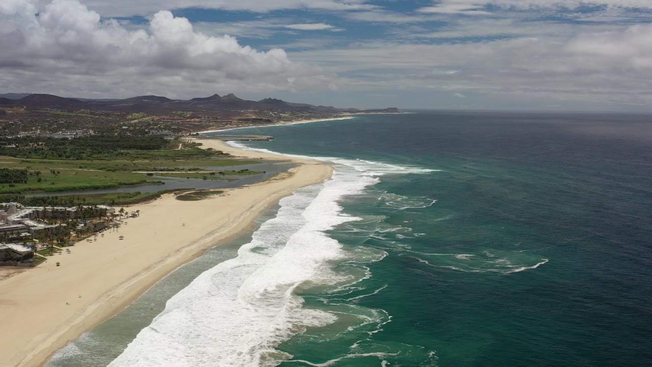 Aerial view over Los Cabos, Mexico showing a long sandy beach, white surf lines, green coastal plain, distant hills, and no visible people at the start of the clip.