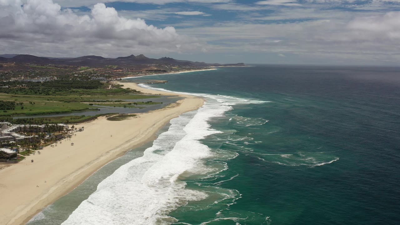Coastal aerial with beach, surf foam, ocean, inland vegetation, and hills.