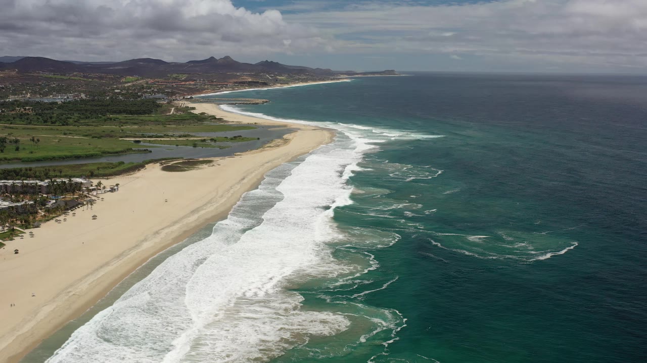 Aerial Los Cabos coastline view with the surf band running diagonally through the frame, pale sand on the left, deep ocean on the right, inland water behind the beach, and no visible people.