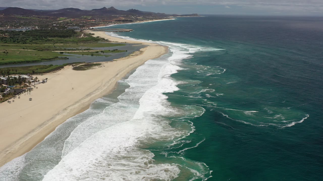 Opening aerial frame over a Los Cabos coastline, with white waves rolling onto a broad tan beach, green land behind it, distant hills on the horizon, and no visible people.