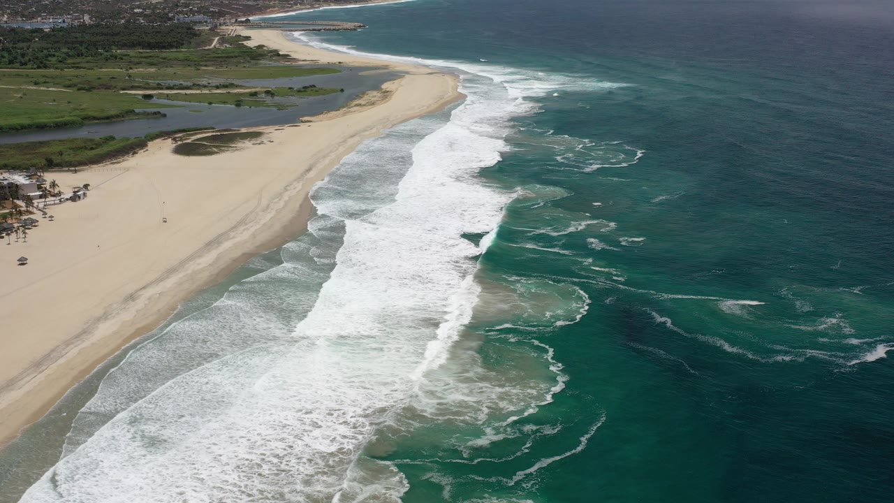 Aerial view over a Los Cabos beach as white surf rolls diagonally across pale sand beside deep turquoise water, with low green shoreline in the distance and no visible people.