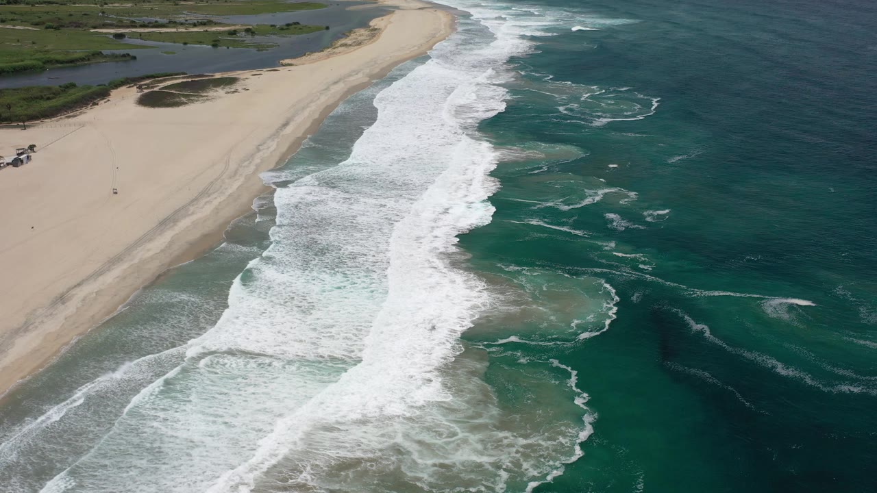 Aerial frame from Los Cabos, Mexico, showing foamy surf bands closer to the lower edge, a broad sand strip on the left, darker ocean water on the right, and no visible people.