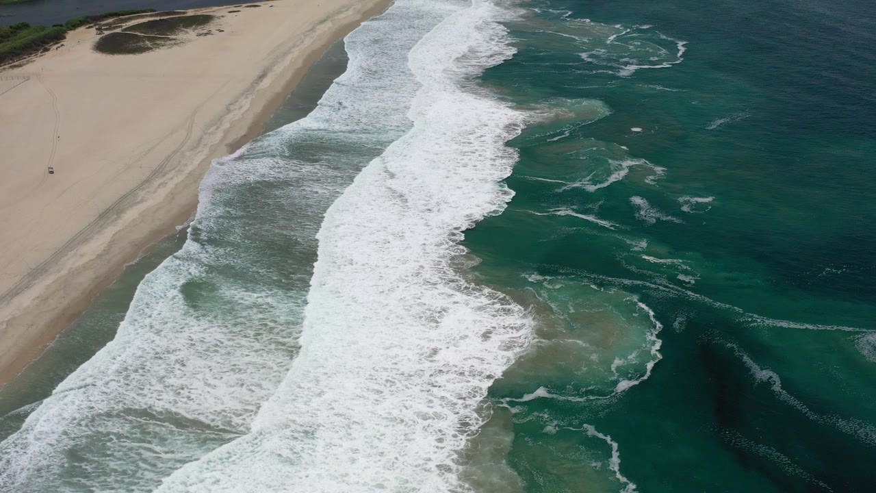 High aerial view of Los Cabos surf breaking in a broad white band between pale sand and dark green ocean, with no visible people.