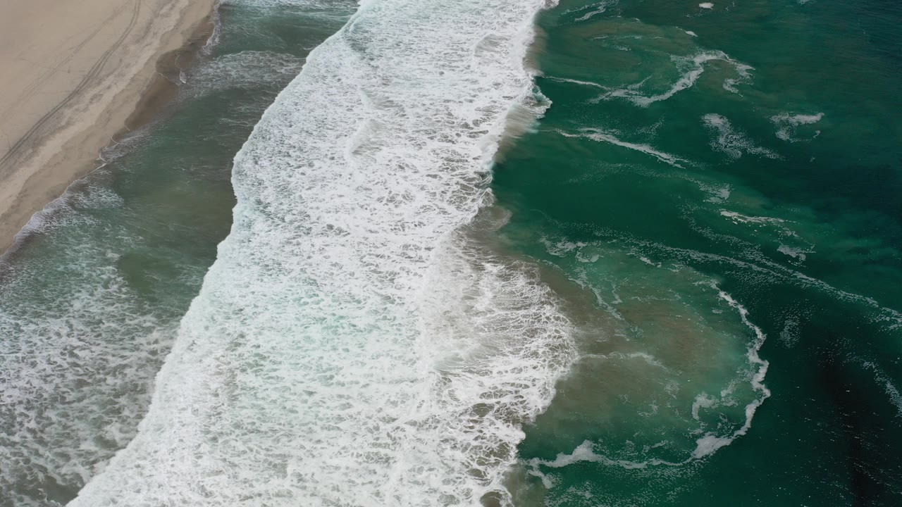 Aerial view over Los Cabos, Mexico with white surf running diagonally beside a tan sandbar and green-blue water, no visible people.