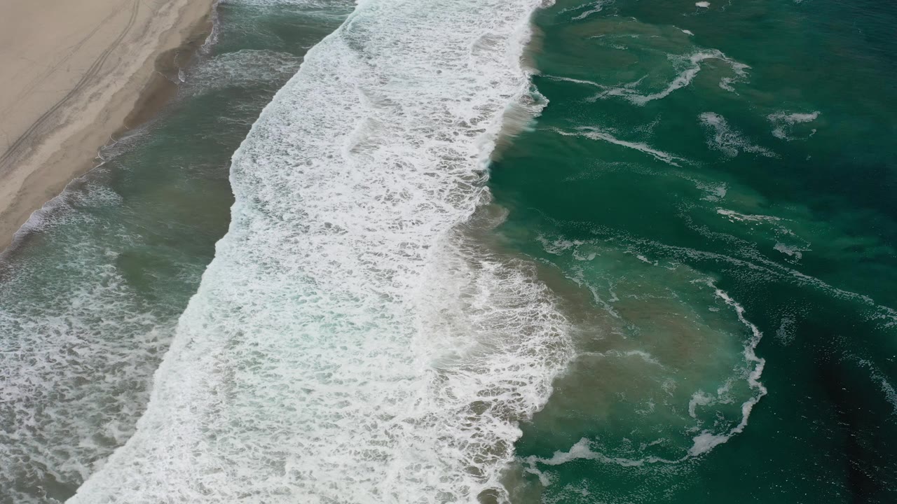 Aerial Los Cabos coastline frame with a broad whitewater band over shallow green water and the sandbar held along the left edge, no visible people.