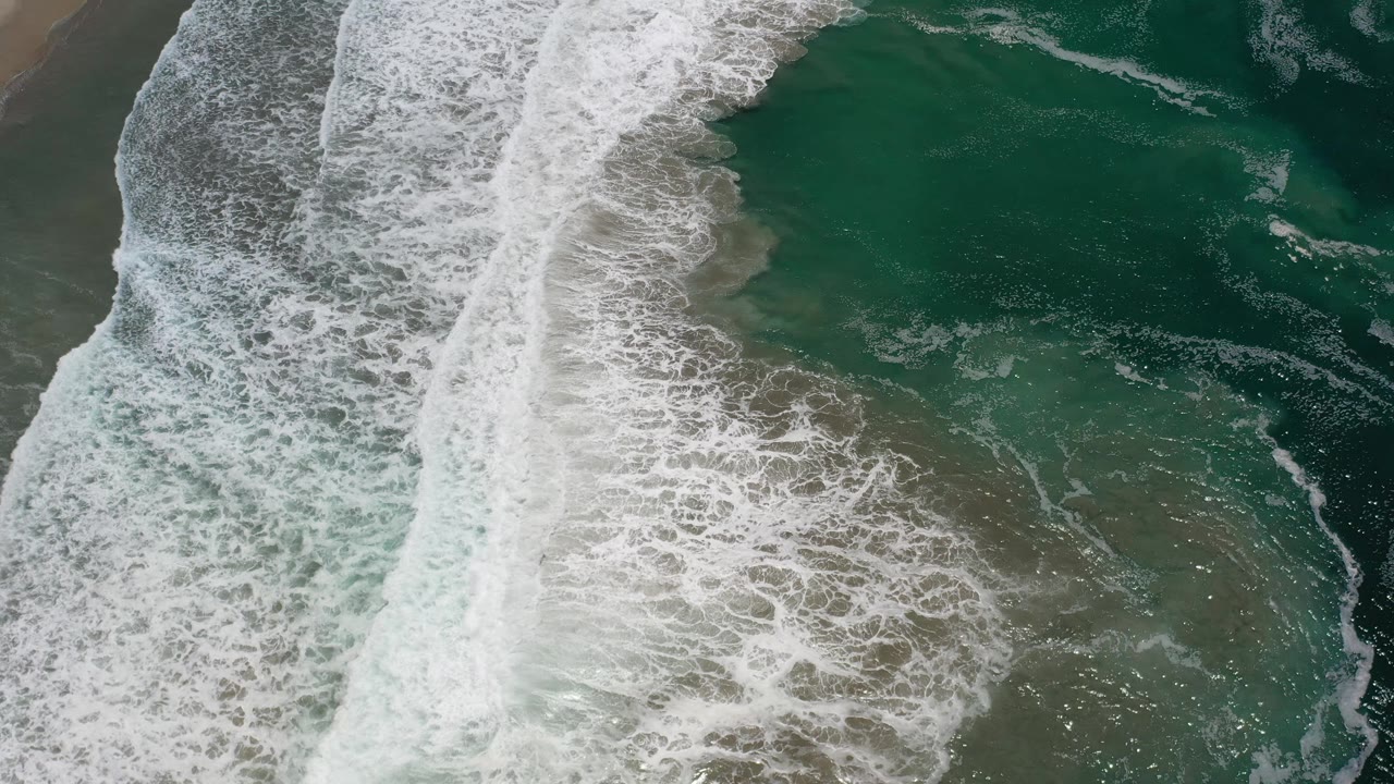 Whitewater spreads wider across shallow green ocean water near Los Cabos, Mexico, while the sandy edge stays partly visible and no people appear.