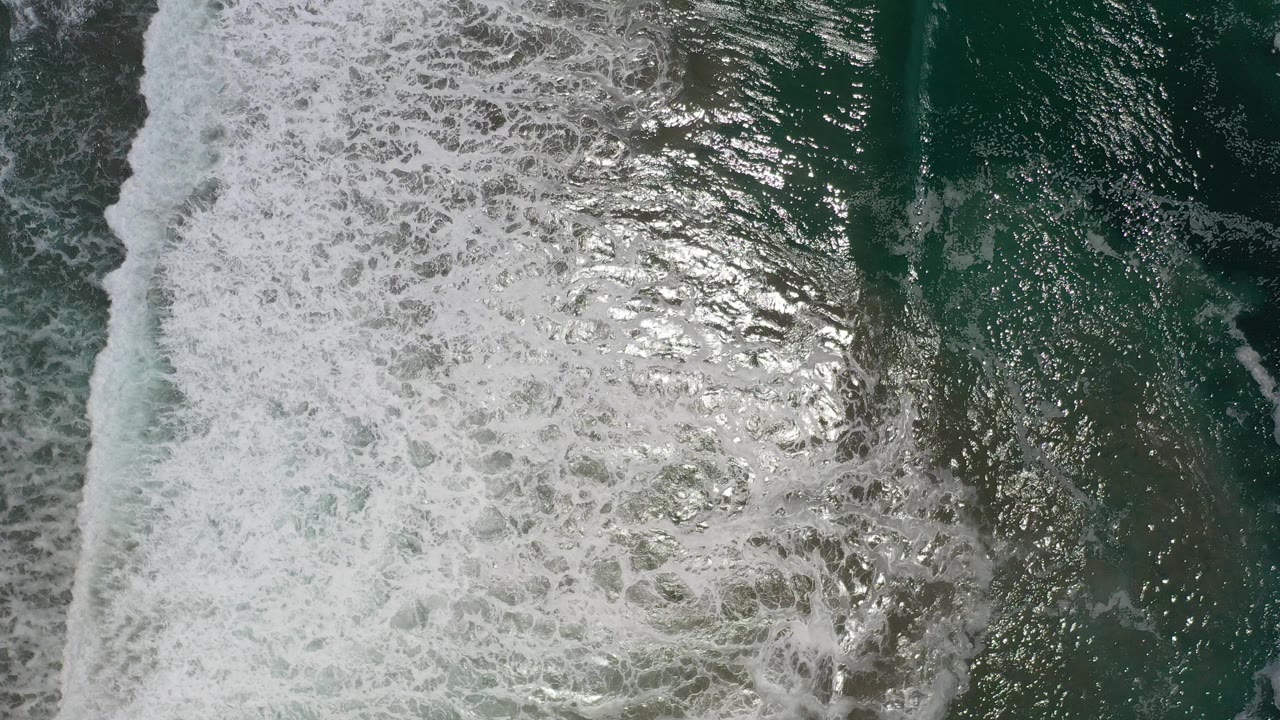 Overhead view of white surf foam meeting dark teal ocean water in Los Cabos, Mexico, with bright surface glare and no visible people.