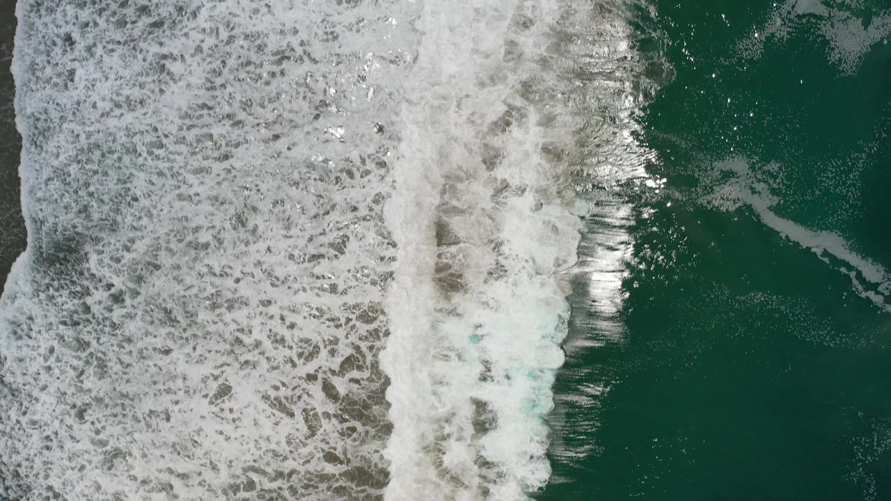 Overhead view of white surf breaking along green-blue water in Los Cabos, Mexico, with wet sand under foam and no visible people.