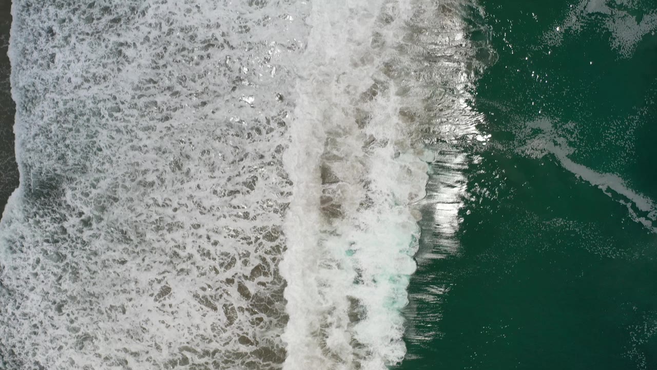 A broad band of foamy white water runs through the frame beside darker green sea in Los Cabos, Mexico, with no visible people.