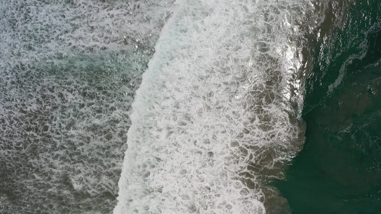 A broad white wave break fills the left side while darker green water and mottled seabed texture sit to the right in Los Cabos, Mexico, with no visible people.