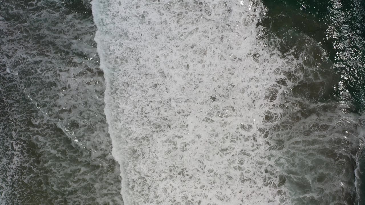 A dense sheet of white foam runs vertically through the overhead Los Cabos ocean view, bordered by darker green water and no visible people.