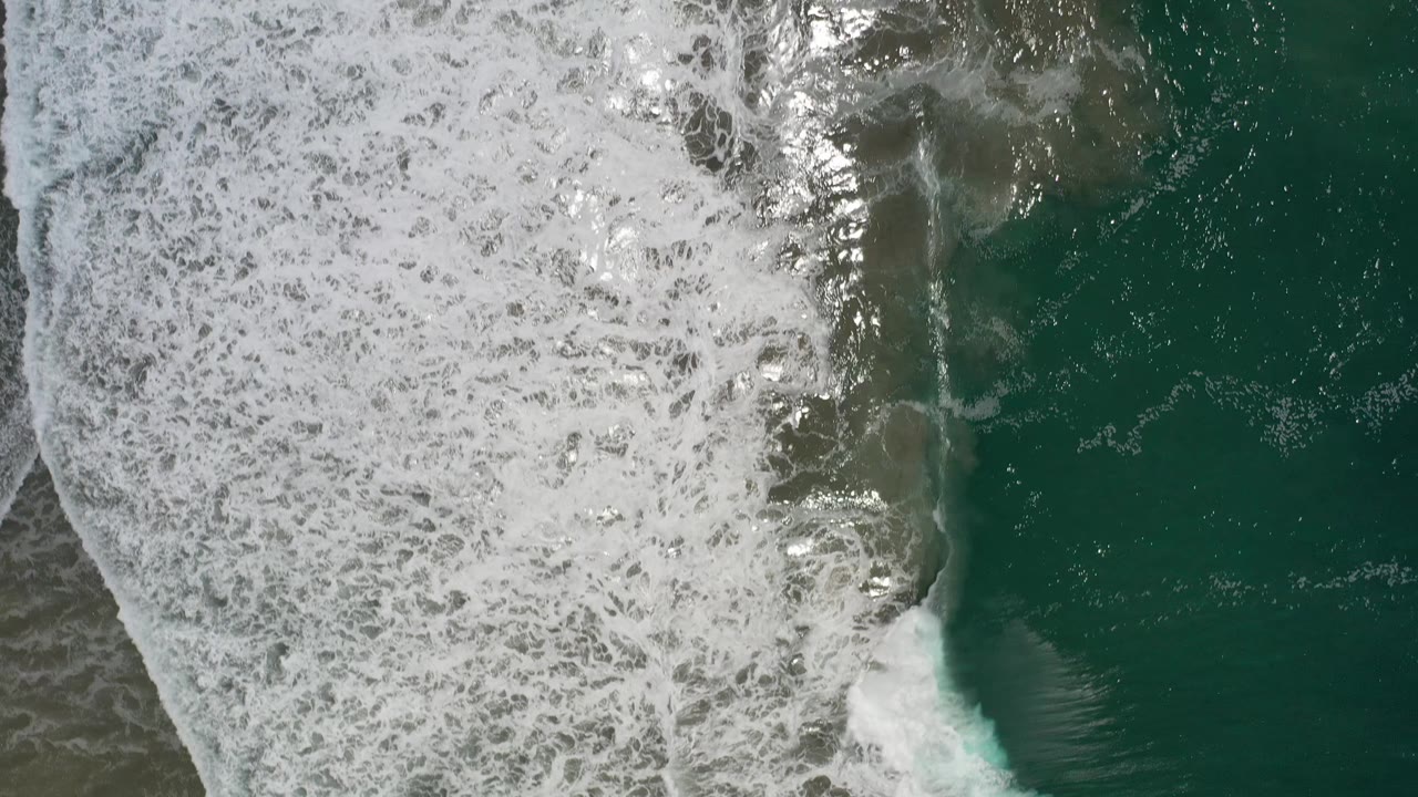 Overhead Los Cabos ocean frame with a thick white foam field on the left and darker green water on the right, with no visible people.