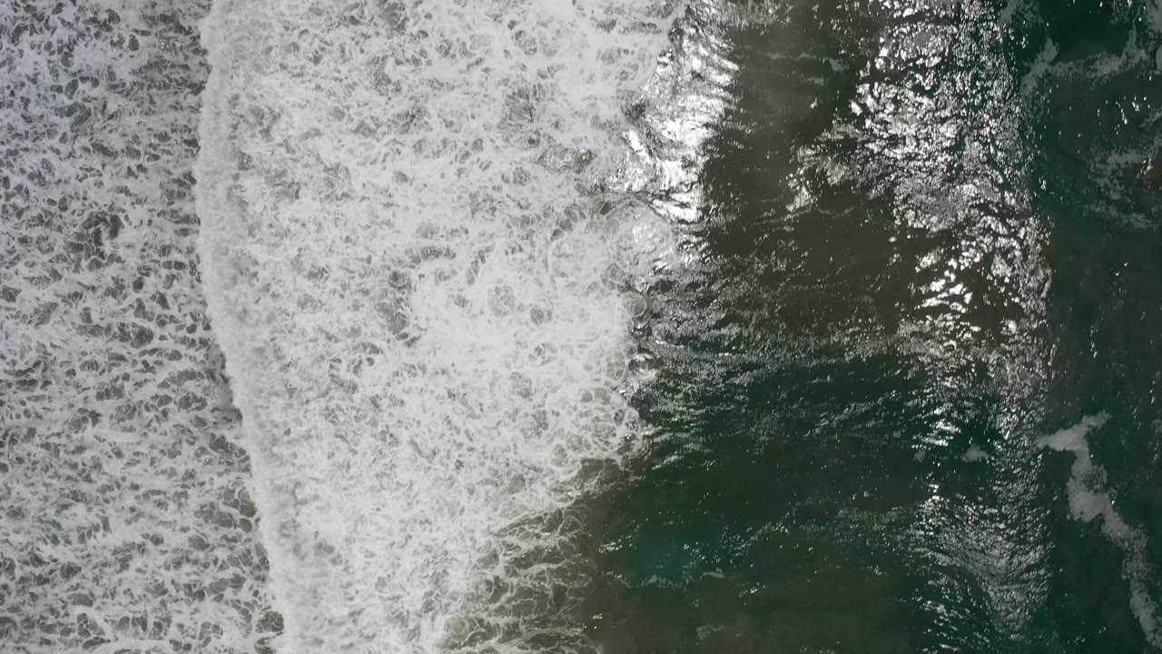 White surf spreads across the left side of the overhead Los Cabos ocean frame while darker green water holds on the right, with no visible people.