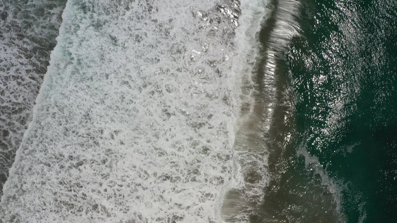 Overhead view of foamy surf meeting green water and a narrow sandy edge in Los Cabos, Mexico, with no visible people.