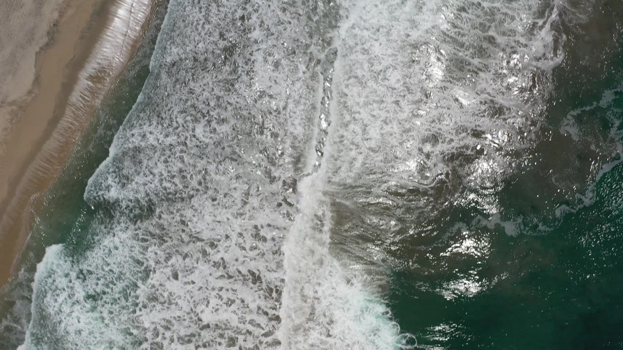 Overhead view of white surf breaking beside wet tan sand in Los Cabos, Mexico, with sun glints on green water and no visible people.