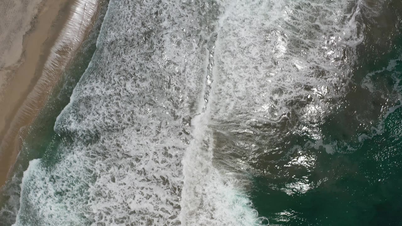 Whitewater spreads farther across green seawater next to a narrow strip of wet sand in Los Cabos, Mexico, with no visible people.