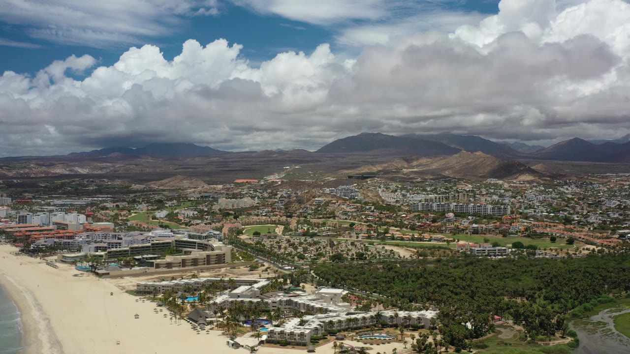Aerial view of beach, coastal buildings, vegetation, hills, and clouds.