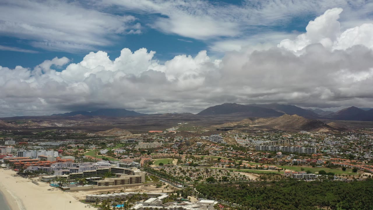 Early Los Cabos aerial frame with resort-scale buildings and streets in the lower frame, a spread of neighborhoods across the valley, brown hills beyond, and no visible people.