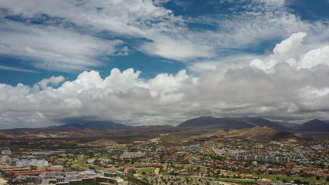 Opening frame of Los Cabos, Mexico from above, with low buildings along the bottom, brown hills and dark mountains across the horizon, large white clouds overhead, and no visible people.