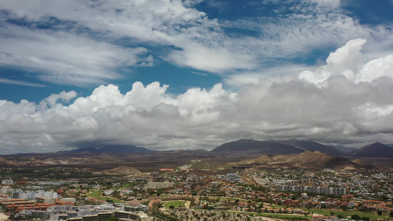 Los Cabos aerial frame with clustered buildings and roads in the lower edge, arid hills in the middle distance, dark mountain ridges beyond, layered white clouds, and no visible people.
