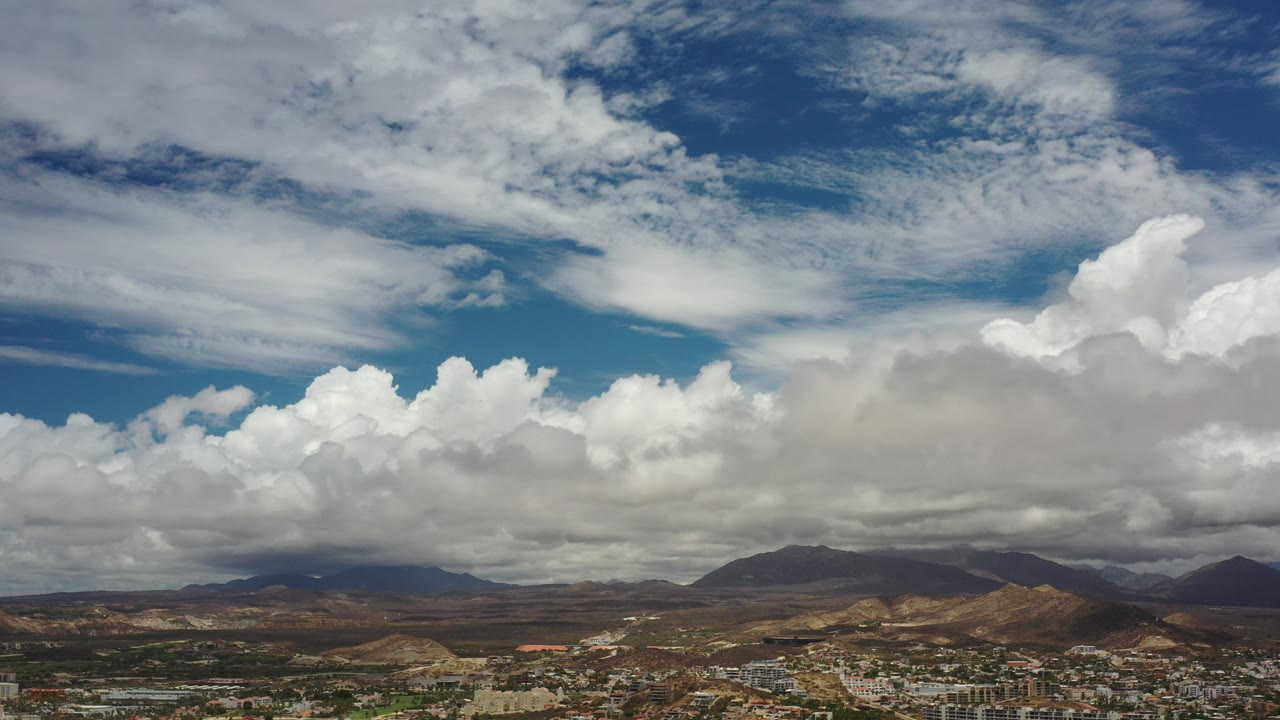 Wide elevated view in Los Cabos, Mexico with layered white clouds over desert hills, distant mountains, a thin strip of buildings, and no visible people.