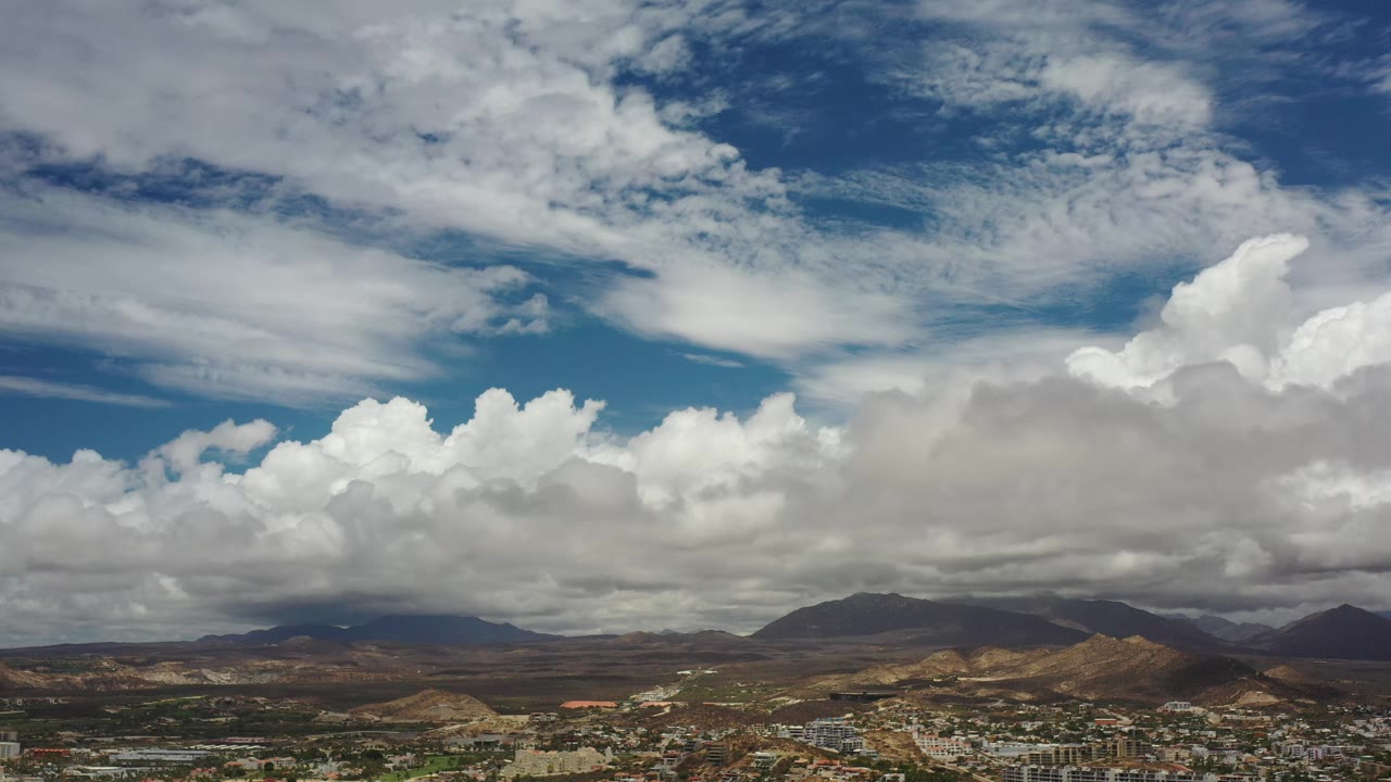 Los Cabos landscape frame with bright cumulus clouds, darker cloud shade over the mountains, brown hills, scattered buildings, and no visible people.