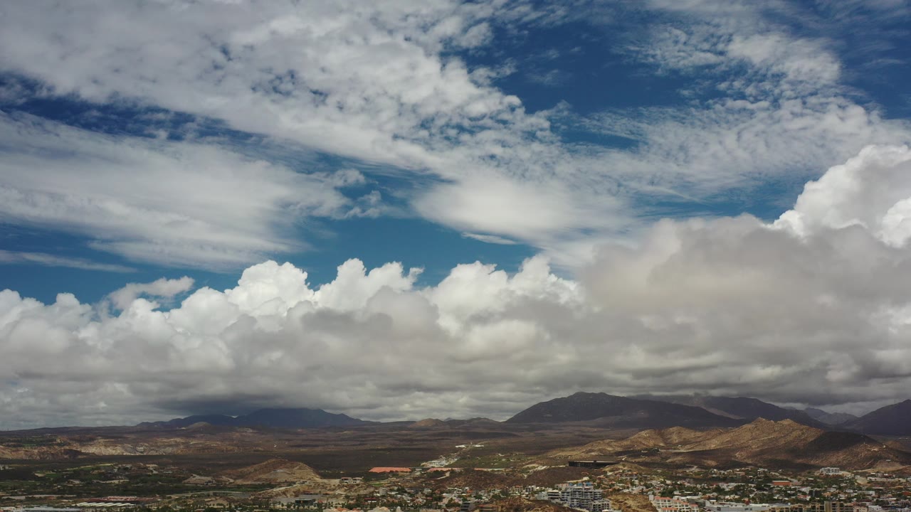Wide Los Cabos, Mexico aerial view with white clouds, dark mountain ridges, dry hills, and a low town along the bottom edge, with no visible people.